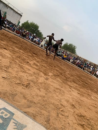 The image shows a wrestling match taking place on a sandy field in Sokoto, Nigeria, located at latitude 13.049 and longitude 5.238. Two men, shirtless, are engaged in a wrestling match. They are in the middle of the field, surrounded by a crowd of spectators. The spectators are seated on the perimeter of the field, with a white building in the background. The men are both wearing shorts and appear to be of African descent. One man is wearing a black pair of shorts while the other is wearing a pair of patterned shorts. The image is taken from a low angle, looking up at the wrestlers. The sand is brown and dry.