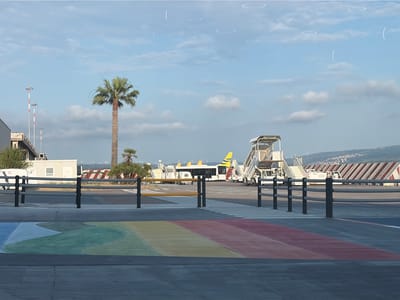 This image captures an airport tarmac scene in Lamezia Terme, Italy, under a clear, bright sky with scattered clouds. The time of day appears to be morning or late afternoon, given the soft light. In the foreground, a vibrant rainbow-colored crosswalk is painted on the asphalt, a striking contrast to the otherwise functional airport environment.

Several airport vehicles and structures are visible. To the left, a white portable building stands near a fence. Further back, a tall palm tree dominates the left-center of the frame, its fronds reaching towards the sky. Behind it, a small cluster of other palm trees can be seen.

The middle ground features airport equipment. A row of metal railings with black posts separates the crosswalk from the tarmac. Beyond the railings, an airport shuttle bus with yellow accents on its tail is parked. Adjacent to the bus are mobile boarding stairs. Further in the distance, more airport infrastructure, including hangars and other vehicles, are present.

The background displays a hilly landscape with buildings and what appear to be wind turbines on the horizon. A red and white striped structure, possibly a hangar or terminal building, is also visible in the distance on the right. The overall scene suggests a typical day at a regional airport, with a touch of artistic flair introduced by the rainbow crosswalk. No people are prominently featured in the image.
