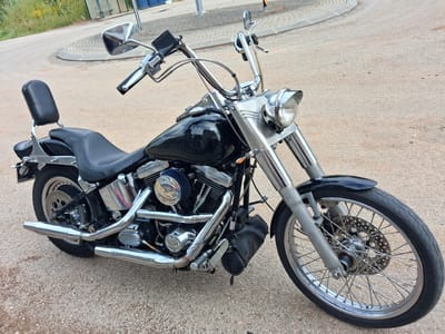 The image shows a black Harley-Davidson motorcycle parked on an asphalt road. The motorcycle features chrome detailing, including exhaust pipes, engine components, and handlebars. A black leather seat with a backrest is visible. The front wheel has spokes. The background includes a roundabout and a building. The image was taken in the city of Soure, Portugal.