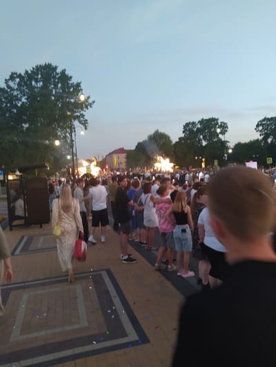 The image depicts a crowd of people gathered on a paved walkway in Gusev, Russia. The sky is overcast with a pale blue hue, suggesting either early morning or late evening. Trees line the background, and streetlights are visible, indicating an urban setting. A bus stop shelter is present on the left side of the image, with a few individuals seated inside. The walkway is decorated with a pattern of squares and is scattered with confetti. Many people are standing, some appearing to be in conversation or watching something in the distance, where a bright light installation is visible. A woman in a light-colored dress is walking away from the camera, carrying a red and white bag. The overall atmosphere appears to be one of a public event or gathering.