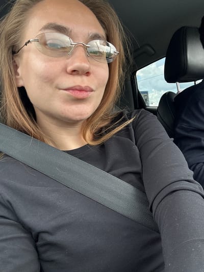 A young Caucasian woman with light brown hair is seen from the chest up, sitting in the passenger seat of a car. She is wearing rimless glasses and a black long-sleeved shirt. A seatbelt is fastened across her chest. Her expression is neutral, and her gaze is directed slightly downwards and to the left. The background shows the interior of the car, including a black headrest of the seat behind her and a window with a view of the sky and some distant structures, possibly indicating an urban or semi-urban environment. The image appears to be a selfie taken inside the vehicle. The exact location in Ardatov, Russia, cannot be determined from the visual information provided in the image.