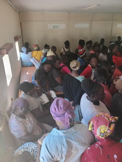 This image captures a large group of people, primarily adults and predominantly women, gathered indoors in what appears to be a community hall or meeting room in Butali Chegulo ward, Kenya. The individuals are seated closely together on benches or chairs, arranged in rows, and are largely facing an unseen focal point, suggesting they are attending a meeting, seminar, or communal event.

Many of the women are wearing head coverings, including colorful scarves and wraps, while others display braided hairstyles. The attire varies in color and pattern, contributing to a vibrant visual. Some individuals are looking towards the front, indicative of listening or engagement, while a few are looking directly towards the camera. The general expressions appear neutral or engaged.

The setting features light-colored walls, possibly cream or beige. A notable detail is a white rectangular sign on the left wall with the text "SEC." clearly visible. In the foreground, there are wooden furniture pieces, possibly benches or tables. The lighting in the room appears adequate, but the time of day or specific weather conditions are not discernible from the indoor setting. The atmosphere suggests an organized community gathering.