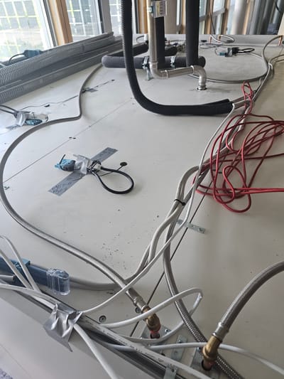 The image shows a close-up, angled view of various pipes and electrical cables attached to a white, textured surface, possibly a ceiling or wall. The scene is cluttered with plumbing and wiring, suggesting an installation or maintenance area.

Prominently featured are several lengths of silver, braided flexible hoses, some connected to brass fittings. Thick black pipes, insulated with black foam, are also visible, joined by silver fittings and secured with clamps. A corrugated black pipe is also present.

Numerous electrical cables, both red and white, are scattered across the surface, some neatly bundled with zip ties, others loosely draped. A small black device, possibly a sensor, is attached to the white surface with silver tape and labeled with handwritten notes. Another small, blue device with a clear plastic cover is also visible among the wires.

The background shows a window with a grid pattern and some greenery outside, indicating an indoor setting with natural light. The overall impression is one of functional, possibly temporary, arrangement of utility lines. No people are visible, and the image is likely taken during daylight hours. No specific time of day, weather, or emotional cues are discernible.

There is some visible text on the white surface and on the cables. One cable has text that appears to read "ISO/IEC 11801 & EN50173..." and another mentions "CAT 6". The handwritten labels on the taped device are difficult to decipher but seem to contain alphanumeric characters and possibly dates.