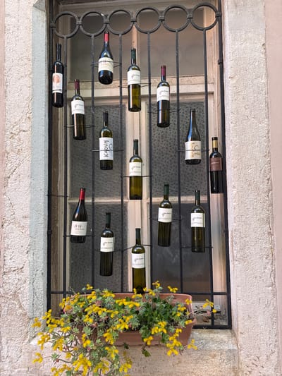 Here is a detailed description of the image:
**General Description:**
The image shows a window in what appears to be a building located in Grad Rovinj, Croatia. The window is barred with a decorative metal grill, upon which a collection of wine bottles are horizontally displayed, attached by thin wires. There is a flower box on the right side of the window filled with yellow flowers, spilling out from behind the bars.
**Architectural Details and Location:**
* **Building Exterior:** The building's exterior wall surrounding the window is made of textured, light-colored stone, consistent with the local architectural style of Grad Rovinj.
* **Window:** The window is composed of small panes of glass.
* **Metal Grill:** The grill design is primarily straight horizontal bars, except for the left side which incorporates decorative circles.
**Objects:**
* **Wine Bottles:** A collection of approximately twelve wine bottles of varying sizes and types are horizontally suspended on the grill. The bottles are suspended with thin wire. The labels on the bottles are readable in some instances, revealing names like “San Zvanini”, "KADUM", "Terra Rossa", "FESTIGIA"
* **Flower Box:** A terracotta-colored flower box is mounted to the right side of the window.
* **Flowers:** The flower box is filled with vibrant yellow flowers, possibly Sedum or similar species, with green foliage.
**Specific details of interest:**
* Some of the bottles are filled, while others might be empty, or only partially filled.
* The arrangement of the bottles appears deliberate, but it's not entirely clear if it serves a functional purpose or if it's solely for decorative purposes.
**Environment:**
The overall scene suggests a warm, Mediterranean climate, characteristic of Grad Rovinj. The building seems to be located on a street or public space, though no people are visible in the image.