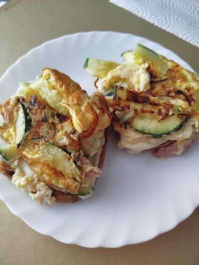Two savory open-faced sandwiches are presented on a white plate, which rests on a neutral-toned textured surface. Each sandwich appears to be built on a base that resembles toasted bread, topped with a fluffy scrambled egg mixture, slices of zucchini, and some pieces of ham. The eggs are golden brown in places, indicating they have been cooked, and the zucchini slices are visible, some with their green skin still attached. The sandwiches are roughly square in shape and are positioned side-by-side on the plate. The lighting in the image suggests an indoor setting, possibly a kitchen or dining area. The background shows a white paper towel with a swirled pattern. There are no people or animals present in the image. The scene is static and appears to be a still life of food. The specific location in Offenbach am Main, Germany, cannot be determined from the visual information provided in the image.