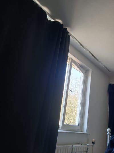 This image depicts an interior residential room located in Oosterhout, Netherlands. The scene is dominated by a partially open window and dark curtains.
On the left, a dark-colored, likely navy or black, curtain hangs from a light-colored rail mounted on the ceiling. The curtain is drawn partially to the left, obscuring a significant portion of the room's left side. Light reflects off the top edge of the curtain where it meets the ceiling.
A white-framed window is visible in the center-right, appearing to be a tilt-and-turn style, slightly ajar inward from its top-left corner. A metallic handle is visible on the left side of the open window pane. Through the window, the outdoor environment shows numerous bare trees with light-colored branches, indicating a leafless season. The sky visible through the window is bright, suggesting daylight.
The wall surrounding the window and extending to the right is painted white. The ceiling is also white, featuring a slight slope, suggesting an upper floor or attic room. A thin, light-colored curtain rail extends across the ceiling from the upper left.
Below the window, a white panel radiator is installed, with a white thermostatic valve visible on its right side. To the far right, a portion of a white, decorative bed frame, possibly metal, is visible with a finial, and some dark fabric, likely bedding, is draped over it.
No individuals are present in the image. The room is illuminated by natural light entering through the open window.