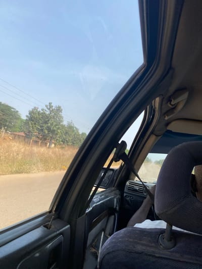 The view from inside a car, angled towards the passenger window. The headrest of the front seat is visible on the right. A seatbelt stretches diagonally across the frame. Outside the window, a bright, clear sky overlooks a landscape of trees and dry grass bordering a road. The car's interior includes the dashboard and door frame. The light is bright and appears to be outdoors.