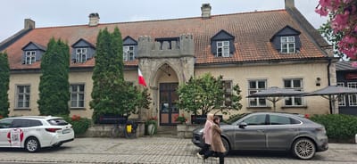 The image depicts the Arensburg Boutique Hotel & Spa in Kuressaare, Estonia. A pale yellow, two-story building with a red tile roof dominates the scene. The building features multiple dormer windows with dark gray roofs and white-trimmed openings. The entrance of the hotel is a large arched doorway framed by stone architectural details, with a flag of Italy hanging from the facade.
In the foreground, a white station wagon is parked to the left, displaying stickers and signage. A dark gray SUV is parked to the right of the station wagon, partially obscuring the building's lower section. Two women are walking in front of the SUV, one in a light pink coat and the other in a brown coat.
A bicycle with yellow saddlebags is parked near the entrance. The ground is paved with cobblestones. Several evergreen trees and bushes are planted around the building, with red flowers visible in planters. Two large dark gray umbrellas are set up in front of a section of the building that appears to be a restaurant or lounge area, indicated by a red sign reading "Restor..." and "Lounge." Pink flowers hang from above on the right side of the image. The sky is overcast.