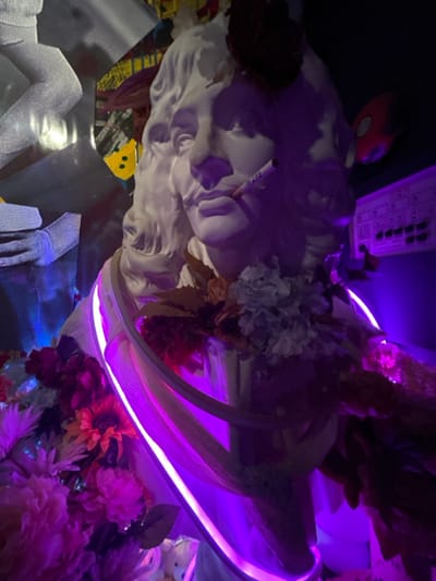 The image displays a white plaster bust of a man with a mustache and wavy hair, positioned with a cigarette, labeled "DUNHIL," inserted into its mouth. The bust is heavily decorated with artificial flowers in various hues including red, pink, orange, and white, intermingled with foliage, draped around its neck and shoulders. A luminous purple LED strip is wrapped around the upper chest of the bust. The scene is under dim, ambient lighting dominated by purple and blue tones, casting pronounced shadows. To the immediate left, distorted reflections appear on fragmented, reflective metallic surfaces. On the right side of the background, a control panel with multiple knobs and indicators is faintly visible against a dark backdrop, subtly illuminated by the purple light, along with a partially obscured red spherical object. This art installation is located within 마포구, South Korea.