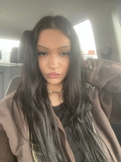 The image displays a female individual seated inside a vehicle in Taverny, France. She has long, dark, wavy hair parted in the middle, extending over her shoulders. Her face features a silver barbell piercing through her left eyebrow and a silver horseshoe-shaped ring in her septum. She is wearing a choker-style necklace composed of multiple light and dark beads. Her lips are colored with a brownish-red tint, and her eyes show dark eyeliner and mascara. She is dressed in a dark brown, possibly fleece-lined, jacket worn over a dark garment. The jacket exhibits a visible snap button closure on the upper right side. Her left hand is partially visible, with fingers appearing near the collar of her jacket. The background, viewed through the vehicle's windows, shows a blurry outdoor environment under what appears to be an overcast sky. A dark car seat headrest is visible behind her on the left.