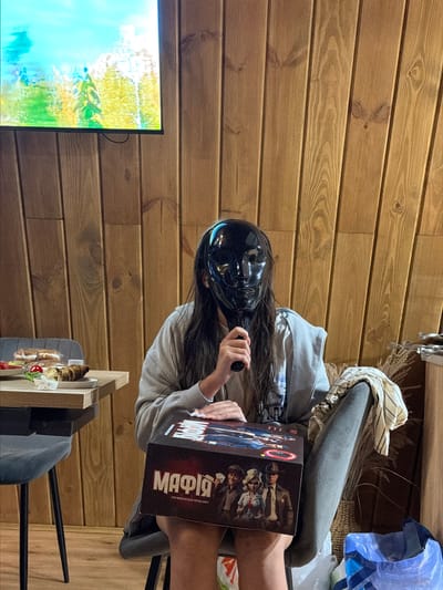 The image depicts a person wearing a black mask sitting at a table in Sharovechka, Ukraine, with a board game box labeled "MAFIA" on their lap. The person is holding the mask up with their right hand. In the background, a television screen displays a nature scene, and the walls are paneled with wood. A partially eaten meal is visible on the table. The environment suggests a casual indoor gathering.