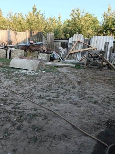 The image depicts a cluttered outdoor scene, likely a yard or storage area, in Leușeni, Moldova. A large, half-cylindrical concrete object lies on the ground in the foreground, partially covered by a thin layer of dirt and sparse vegetation. A long, flexible hose snakes across the ground, extending from the lower left towards the middle right of the frame.
In the background, a pile of assorted materials is haphazardly stacked. This includes various sheets of corrugated roofing material, some leaning against a fence, others supported by wooden beams. A wooden sawhorse-like structure stands among the debris. Interspersed with the building materials are pieces of concrete, metal pipes, and what appears to be tangled wire or mesh. A bright pink helmet rests on a pipe.
Behind the accumulated items, a dense line of green trees forms a backdrop, with a clear, pale blue sky visible above. The lighting suggests it is either early morning or late afternoon, with the sun casting soft shadows. The overall impression is one of disuse and accumulation of discarded items, possibly indicating a renovation project or a junkyard. No people are visible in the image. There is no discernible text in the image.