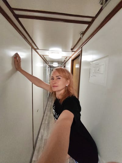 A woman with shoulder-length blonde hair and wearing a black t-shirt leans against a white wall in a narrow hallway. The hallway has white walls, a wooden trim along the top, and a light-colored floor. The woman is looking towards the camera with a slight smile. Her left arm is extended and resting on the wall. She is wearing earrings and has red nail polish. There are lights on the ceiling and a map or diagram is posted on the wall to her right. The image appears to be taken indoors, possibly on a boat or in a building with a similar design. The location of Larionovo, Russia is not visible or suggested by the image.