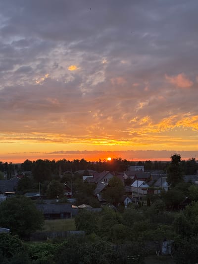 The image captures a panoramic view of a rural settlement at sunset, likely in the Пратасевіцкі сельскі Савет, Belarus. The sky is filled with a dramatic display of warm colors, with the sun low on the horizon, casting a vibrant orange glow across the clouds. The clouds themselves are a mix of grays, purples, and bright oranges, with some illuminated by the setting sun.
Below the sky, a dense line of dark green trees forms a natural border across the middle of the frame. Peeking through and beyond this tree line are the rooftops of various houses, predominantly featuring gray and dark-colored roofing materials. The architecture suggests a traditional rural setting, with houses of varying sizes and styles, some appearing older with weathered roofs and others more modern. Small chimneys and antenna are visible on some structures.
The foreground is dominated by lush green foliage, with trees and bushes creating a dense, natural canopy. Wooden fences, characteristic of rural properties, are visible in some areas, delineating yards and gardens. The overall impression is one of a peaceful, twilight scene in a village surrounded by nature. There are no discernible people or animals in the image. The lighting suggests late evening, with the day transitioning into night.