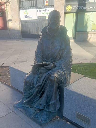 The image shows a bronze statue of a seated man in religious attire, likely a friar, holding a book. The statue is positioned on a grey stone bench in what appears to be a public square or walkway. Behind the statue, there is a building with a large metal gate and a sign that reads "Educación Infantil y..." and another sign for "CLUB DEPORTIVO COLEGIOS DIOCESANOS". The ground in front of the bench is paved with square tiles, and a patch of green grass is visible to the right. The lighting suggests it is daytime, with shadows cast by the statue and the building. A nameplate on the front of the bench reads "TOMÁS LUIS DE VICTORIA". This statue is located in Ávila, Spain, specifically in the Plaza de Tomás Luis de Victoria.