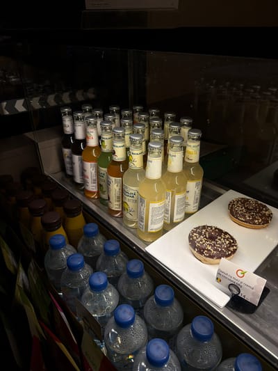 This media file captures a refrigerated display case, likely found in a store or cafe in Florești, Moldova, showcasing an assortment of beverages and a sweet treat.
The prominent subjects are two chocolate-covered donuts and a variety of bottled drinks. The donuts, resting on a white rectangular plate, are coated in dark chocolate and generously topped with chopped nuts. Adjacent to them is a white price tag in Romanian, reading: "Donut cu nuci și cremă de ciocolată 68g" (Donut with nuts and chocolate cream 68g) and "PRETUL 14 lei" (PRICE 14 lei), indicating the local currency.
To the left of the donuts, numerous small glass bottles are neatly stacked, many branded "SICILIANA" and featuring various flavors such as "LIMONATA" (lemonade), "COLA," "LEMON BITTER," and "DI SAMBUCO" (likely elderflower). A dark bottle further left is labeled "CEAI EARL GREY" (Earl Grey Tea), also displaying its Russian/Ukrainian translation "Черный чай Эрл Грай." The bottles appear to be 275ml in volume, with brands like "Bona" and "1947" visible on some labels.
On the right side of the display, several clear plastic bottles of water with blue caps are stacked, showing condensation that suggests they are chilled. Below these are rows of smaller bottles with yellow caps, possibly juices or other drinks. The overall scene depicts a well-organized selection of cold refreshments and snacks available for purchase.