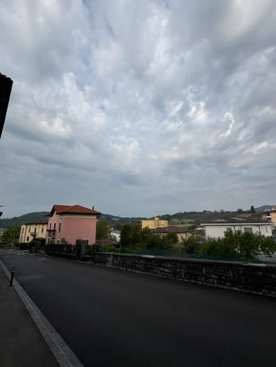 The image depicts a view of a street in Balerna, Switzerland, on a cloudy day. The foreground shows a dark asphalt road with a granite curb and a small black bollard on the left side. Beyond the road, a stone wall separates the street from a residential area. Several houses are visible, including a prominent pink building with a red tiled roof and a yellow building with multiple stories. In the background, rolling hills covered in vegetation and some terraced fields are present. The sky is filled with dramatic, grey clouds, suggesting overcast weather. The overall scene conveys a quiet, suburban or small-town atmosphere. There are no people or animals visible in the image.