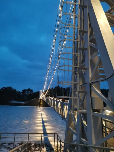 The image displays a pedestrian suspension bridge at dusk or night, located in Мастоўскі сельскі Савет, Belarus. The bridge features white structural towers, one visible prominently on the right with an affixed ladder, and numerous illuminated lights strung along its suspension cables. These lights cast elongated reflections and shadows across the largely frozen body of water below. Several individuals are discernible walking on the bridge's walkway. The background reveals a dark line of evergreen trees along the far bank of the water. The sky is dark blue, characteristic of twilight or nighttime conditions.