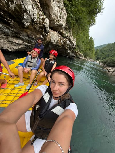 Five people are on a yellow raft, preparing to go whitewater rafting on a river in a rocky canyon. The raft is positioned on the water, with a steep, rocky cliff face covered in lush green vegetation on the left side of the image. The river is a deep, murky green color, with ripples on its surface. The sky is overcast.

The people are wearing helmets and life vests. One woman in the foreground, facing the camera, is taking a selfie with a wide smile. She is wearing a red helmet and a black life vest over a white t-shirt and white shorts. Another woman, to her left and slightly behind, is wearing a blue patterned top and a life vest, holding a paddle. Behind her, a man in a dark blue t-shirt and a red helmet is looking towards the camera. Further back, a woman with a red helmet and black life vest is looking forward. The hands of another person, holding a paddle, are visible in the lower part of the frame.

The overall mood is one of excitement and anticipation for the rafting adventure. The image appears to have been taken during the daytime, given the natural light. The location is Džajići, Bosnia and Herzegovina, indicated by the rocky canyon and dense vegetation typical of the region. There is no visible text in the image.