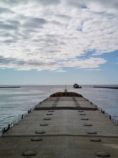 The image presents a view from the deck of a large cargo ship, possibly a bulk carrier, sailing on a body of water. The deck is grey and has numerous circular hatches. The ship is heading towards the open sea, passing between two breakwaters marked by small lighthouses. In the distance, another ship is visible. The sky is partially cloudy, with patches of blue visible. Based on the location, it appears that the vessel is near Ozernoe Ust'e, Russia, as that is the region described in the query. The water is choppy and grey.
