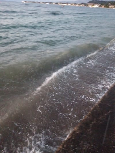 This is a ground-level, medium shot image taken at dusk or dawn on a rocky or paved shoreline in the Municipal Unit of Vrachneika, Greece. The water is dark and choppy, with white foam visible as a wave breaks along the edge of the land. The shoreline itself appears to be made of dark, rough material, possibly concrete or large stones. In the distance, a hazy line of lights suggests a settlement or town along the coast. The sky is overcast and a pale, grayish-blue. No people are visible in the frame.