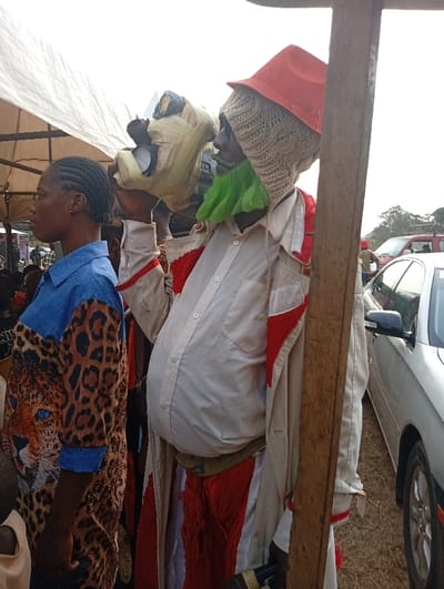 The image depicts a cultural event taking place outdoors in Onicha, Nigeria, likely during the daytime.

The central figure on the right is a person in an elaborate costume, possibly a masquerader. They wear a light-colored, long-sleeved coat with red accents, over a white button-down shirt that appears padded or oversized, creating a bulging stomach. A red, fringed lower garment is visible beneath the coat. On their head, they sport a textured, beige knitted head covering, topped with a bright orange, conical hat. A vibrant green, fringed beard or facial covering obscures part of their face. The individual holds a light-colored, intricately crafted mask or headpiece, possibly made of raffia or woven material, with dark eyeholes, positioning it beside their head. A sturdy wooden post partially frames this figure on the right.

To the left, a woman stands in profile, observing the scene. She has dark skin and her hair is styled in braids. She wears a distinctive shirt featuring a blue upper section and a bold leopard print pattern below, which includes a prominent leopard head with bright blue eyes on the chest.

In the background, a white canvas canopy or tent structure provides shade, indicating an organized gathering. A silver car is parked on the right side of the frame. The scene suggests a communal celebration or a traditional performance. There is no visible text in the image.