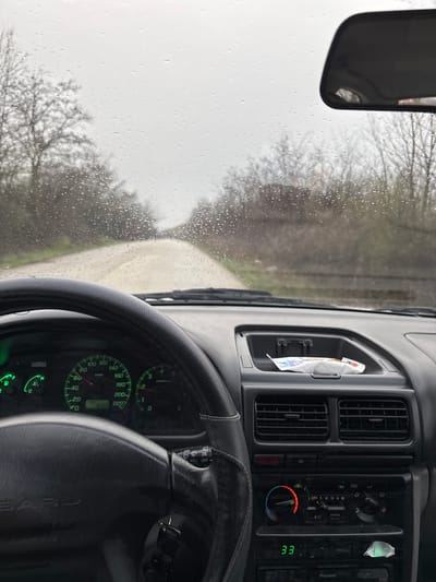 The image depicts the interior perspective from the driver's seat of a vehicle, in the city of Suhache, Bulgaria, looking forward through a rain-splattered windshield. The environment outside shows an unpaved or dirt road, wet from rain, extending into the distance under an overcast sky. Bare trees and sparse vegetation line both sides of the road.
Inside the vehicle, the black steering wheel, bearing a "SUBARU" logo, dominates the lower left quadrant. Behind it, the instrument cluster is partially visible, showing a speedometer indicating approximately 20 km/h and a tachometer around 1000 RPM, along with a fuel gauge, temperature gauge, and an odometer displaying "TRIP 190.2." Keys are visible in the ignition on the right side of the steering column. A wiper control stalk is also visible on the right.
The central dashboard features air vents, a storage compartment containing a white crumpled wrapper or tissue, and climate control dials set to a warm temperature, with the A/C button visible. Below these controls, a digital display shows "33," and a "Pioneer" brand head unit is present. A rearview mirror is partially visible at the top center of the windshield. No people are visible within the frame.