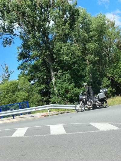 In Strunkovice nad Volyňkou, Czechia, a motorcycle rider wearing a black riding suit and a yellow helmet is positioned on a motorcycle with two large cases mounted on the rear. The motorcycle is on the right side of a paved road, angled slightly towards the left. The road features white lane markings and a pedestrian crossing. A silver guardrail runs along the edge of the road, with a blue fence visible beyond it. The background is dominated by lush green trees and a clear blue sky with some white clouds. The scene appears to be a roadside setting with natural elements.