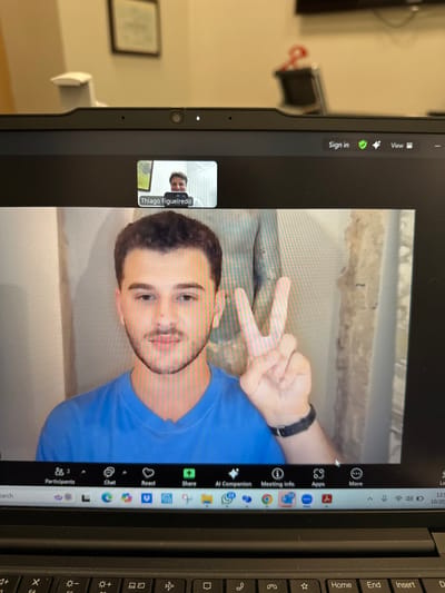 The image depicts a close-up view of an active video call displayed on a laptop screen, situated in an indoor environment within Aventura, United States. The laptop's webcam is active, indicated by a small white light above the screen.
The primary video feed on the screen shows a young male with short dark hair, a light mustache, and a short beard, wearing a solid blue t-shirt. He is smiling directly at the camera and holding his right hand up, forming a "peace" sign with his index and middle fingers extended. A black watch is present on his left wrist. His background consists of a light-colored wall, partially discolored or textured on the right.
In a smaller, inset window at the top center, a second male participant, identified as "Thiago Figueiredo," is visible. He is smiling, with a brightly lit background suggesting an outdoor or well-lit indoor setting with green elements.
The laptop's interface, consistent with a video conferencing application like Microsoft Teams, displays controls at the bottom, including "Participants (2)", "Chat", "React", "Share", "AI Companion", "Meeting info", "Apps", and "More." The system tray on the lower right indicates the time as 12:51 PM and the date as 10/20.
The physical environment behind the laptop includes a light-colored wall, a framed object (possibly a certificate or picture) hanging on it, and partially visible objects such as a chair back on the left and a dark object with a red bow or ribbon on the right.