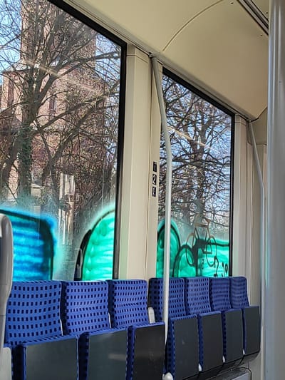 The scene depicts the interior of a passenger vehicle, likely a train or tram, positioned within the city of Einbeck, Germany. The perspective is from inside, looking outwards through multiple windows on a clear, sunny day.
The vehicle's interior foreground features a row of unoccupied blue, checkered fabric seats with dark grey backings, extending horizontally across the lower frame. The interior walls and ceiling are cream-colored, complemented by light grey vertical support poles visible between the windows and towards the right. A small black sign with white symbols, including the numeral '2' and a bicycle icon, is affixed to the cream wall between the left-central windows.
The exterior view through the windows is significantly obscured by graffiti. The lower portions of all visible windows are covered with bright spray paint. The left-most window displays broad strokes of blue graffiti. The central and right windows exhibit vibrant green graffiti composed of rounded, organic shapes. Superimposed over the green graffiti in the right-most window is a black outline drawing of a bicycle, with the numerals "20" visible directly beneath it.
Beyond the graffiti, through the clear upper sections of the windows, numerous bare deciduous tree branches are discernible against a bright blue sky, indicating a late autumn or winter season. Through the upper left window, a large, reddish-brown brick building, consistent with a church or historic structure featuring a prominent tower, is partially visible amidst the tree branches, characteristic of architecture found in Einbeck.