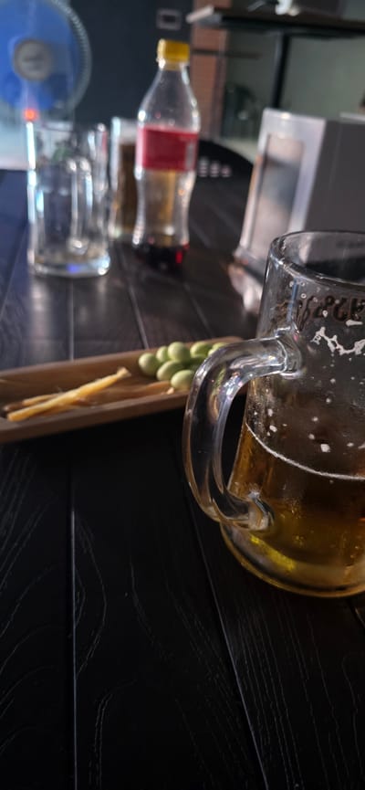 A close-up shot, angled from above and to the side, captures a table setting at what appears to be a casual establishment in Gori, Georgia. The dark, wooden table surface has a textured, wood-grain pattern. In the foreground, a glass beer mug, approximately one-third full with a golden-hued beverage and frothy head, sits to the right. The mug is slightly condensation-covered. To the left of the mug, a small, rectangular wooden tray holds a snack. On the tray are several light green, oval-shaped olives and two thin, yellowish sticks, possibly cheese or dried meat. In the background, slightly out of focus, are several glasses and a red and clear plastic bottle of what appears to be a carbonated beverage. The environment suggests a relaxed, possibly social, gathering.