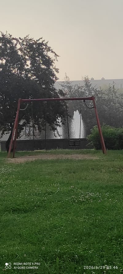 The image depicts a park or playground scene in Maardu, Estonia. In the foreground, a green grassy area with small white flowers is visible. A red metal swing set with two swings, one with a yellow seat and the other with a black seat, stands in the middle ground. Behind the swing set, a fountain sprays water upwards. Trees and foliage surround the fountain, and a building is visible in the background. The sky is overcast, creating a soft, diffused light. The bottom of the image contains a watermark with camera information and a date stamp.