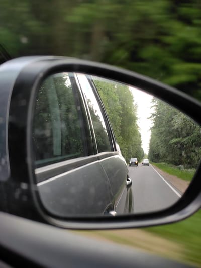 The image shows a reflection in a car's side mirror, depicting a road lined with trees. Two cars are visible on the road ahead, traveling in the same direction as the camera car. The scenery suggests a rural or suburban setting, possibly on a highway. The image does not provide specific details to identify the exact location as Ilyanskiy selskiy Sovet, Belarus, or any other city.