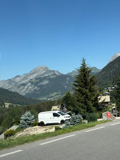 This media file shows a scenic view of a mountain landscape in Châtel, France. A white van is parked on the side of a paved road, which has white dividing lines. The van is positioned on a gravel and grass verge. In the background, a steep, rocky mountain dominates the view, with green slopes dotted with trees and some buildings. The sky is clear and blue, suggesting a sunny day. The time of day appears to be daytime, likely late morning or afternoon, given the bright sunlight casting shadows. The overall mood is serene and picturesque, highlighting the natural beauty of the mountainous region. There is a red cylindrical object with the number "707" visible on a concrete structure by the roadside.