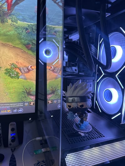 The image shows the interior of a computer, with a gaming screen visible on the left. The computer has multiple illuminated fans, and a Funko Pop figure of Kakashi Hatake from Naruto is placed inside. The scene is illuminated by the blue light of the computer's fans. The location is not specified in the image or context, and therefore cannot be determined to be Castillo Bajo-conejito, Spain.