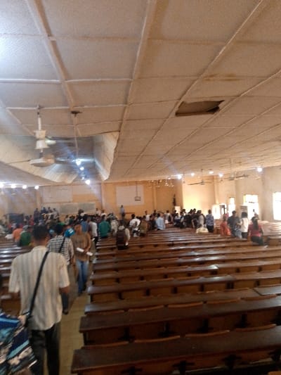 The image depicts the interior of a large academic lecture hall or auditorium, likely situated within a university campus in Alabata, Nigeria.

The room features a suspended panel ceiling exhibiting significant wear, discoloration, and visible water staining. A large section of a ceiling panel or insulation is partially detached and sagging on the left, while a rectangular panel is entirely missing in the upper-right quadrant, exposing an opening to the space above. Multiple ceiling fans with integrated light fixtures are distributed throughout the space, along with recessed lighting. The walls are light-colored, possibly beige or yellow. A large, plain white board or screen is mounted on the far wall, with another white rectangular object visible further to its right.

Numerous rows of dark wooden benches with integrated desks or seating are arranged in an auditorium style, facing the far end of the hall. The foreground rows are largely empty, while the middle and rear sections are occupied by a significant number of individuals, predominantly young adults.

In the immediate foreground, a male subject wearing a light-colored, short-sleeved striped shirt and dark trousers, carrying a dark shoulder bag, walks away from the camera in a central aisle. To his right, another male subject in a light brown shirt and blue jeans walks towards the camera in an adjacent aisle, holding a white document or paper in his hand. Further back, individuals are observed seated on the benches, standing, and congregating in the aisles, with some appearing to face the front of the hall. Natural light enters from windows visible on the right wall, complementing the artificial lighting from the ceiling fixtures.