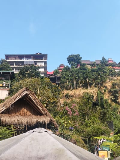 The image depicts a lively outdoor scene with a clear blue sky, suggesting a sunny day, likely during daylight hours. In the foreground, the curved, light-colored surface of a large canopy or tent dominates the lower part of the frame. Behind it, a traditional thatched-roof hut with a steep pitch is visible, providing a rustic contrast to the more modern structures.

The mid-ground is a lush, green hillside covered in dense vegetation and trees, interspersed with buildings. On the left, a multi-story building with a white facade and dark windows is partially obscured by trees. Further up the hillside, a larger building with distinct, multi-tiered red roofs and white accents is visible, showcasing a traditional architectural style.

In the background, more of the hillside is covered in trees, with some bare, dry branches adding texture to the landscape. Along the right side of the image, a set of stairs or a pathway is lined with colorful signs and people. The presence of multiple people, some walking up the stairs and others gathered near the signs, suggests a public space or tourist attraction.

One sign is a bright yellow with red text that appears to read "PULAU GOREE" and "SHINTODOKU RYOKEN". Another green sign has white text. The overall impression is one of a vibrant, possibly tropical or mountainous location in Indonesia, with a blend of natural beauty and architectural elements, bustling with people. The atmosphere appears energetic and inviting, with no indications of negative emotions.