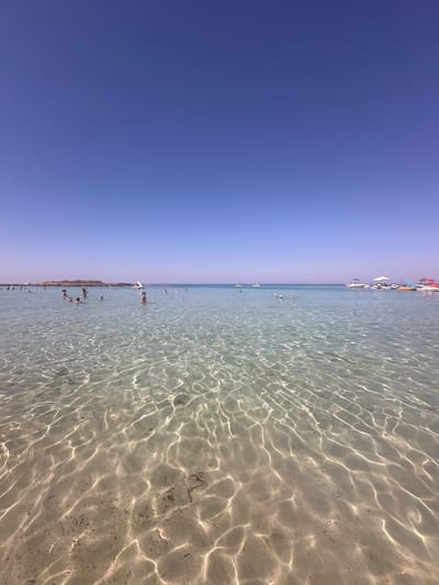 The image depicts a clear, shallow sea with a sandy bottom, under a bright blue sky. The water is calm, with sunlight creating shimmering patterns on the seabed. Several people are visible in the water, some swimming and others standing. In the background, there are boats docked near the shore and a distant island or landmass. The scene is consistent with a popular beach destination. This image is likely taken at Fig Tree Bay in Protaras, Cyprus, a well-known beach celebrated for its shallow, clear waters and golden sand.