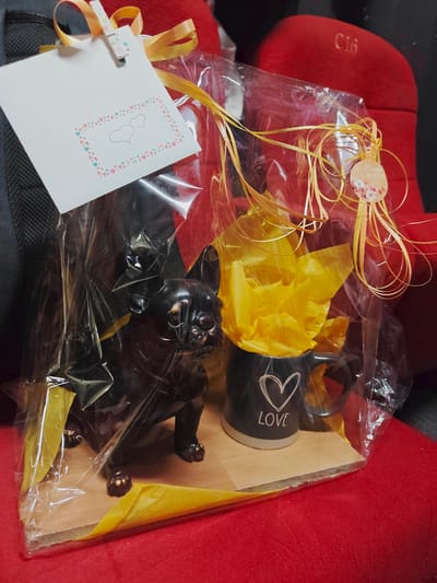 A gift-wrapped item is displayed on a red surface. The gift includes a dark-colored French bulldog figurine and a black mug with a heart and the word "LOVE" printed on it, set on a wooden base. The items are enclosed in clear plastic wrapping, adorned with yellow ribbon and a small tag that reads "Plaisir d'offrir". A white card with a heart design is attached to the wrapping with a decorative clothespin. In the background, red theater seats are visible, with one seat marked with "C16". The scene appears to be in the city of Kirrwiller, France, likely within a theater or performance venue.