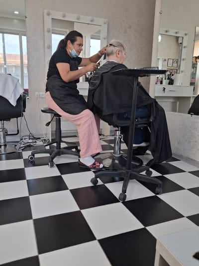 A woman with her hair in a ponytail, wearing a black t-shirt, a black apron, pink pants, and floral-patterned slip-on shoes, is seated on a black rolling stool. She is masked and using scissors and a comb to cut the hair of another person, who is wearing a black cape. This person is seated in a chair and has short, blonde hair. The scene takes place inside a brightly lit hair salon. The floor is tiled with a black and white checkerboard pattern. In the background, there are mirrors with lights around them, shelves with various hair products, and windows that look out onto a building and some trees. The setting appears to be a commercial establishment within the city of Tuzdybastau, Kazakhstan.