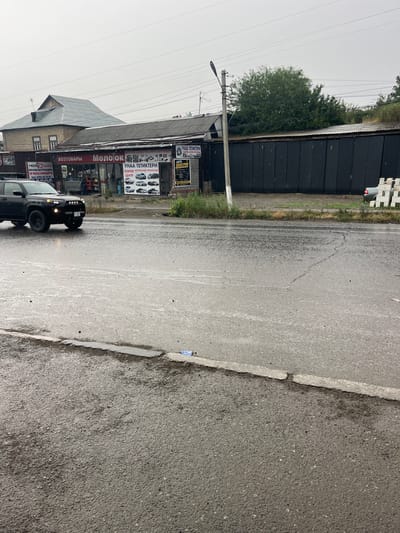 The image captures a street scene in Medrese, Kyrgyzstan, likely on a rainy or overcast day given the wet asphalt and muted lighting. The foreground is dominated by a wet, grey asphalt road with visible cracks and a raised curb on the left. In the mid-ground, a black SUV is driving on the wet road, its headlights illuminated. To the right of the road, a row of buildings lines the street. One building, likely a commercial establishment, features signage with Russian text, including "ХОЗТОВАРЫ" (Household Goods), "МОЛОКО" (Milk), and "УНА ТЕТИКТЕРИ" (possibly a service related to car parts, with an image of cars). Another sign indicates "АВТО ЭЛЕКТРИК" (Auto Electrician). Behind these structures, a dark corrugated metal fence stretches across the background, obscuring further details. Green foliage is visible above the fence and to the left of the buildings. A single streetlight stands on a pole in the mid-ground. The sky is a uniform, overcast grey.