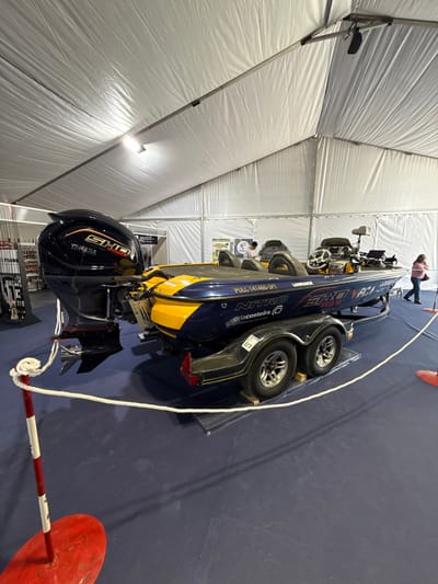 The image depicts an indoor exhibition scene, likely an event or trade show, located in the city of Mora, Portugal. The primary subject is a dark blue and yellow bass fishing boat, branded "NITRO" and "arlacosteira," displayed on a dual-axle trailer. The boat is equipped with a black "YAMAHA SHO VMAX" outboard engine at the stern and features "LOWRANCE" electronics at the steering console. The trailer wheels rest on wooden blocks positioned on dark gray ramps, with a white sticker bearing "07" in a green circle on the fender. The boat is surrounded by a white rope barrier, supported by red and white striped poles with red circular bases, demarcating the display area on a dark blue carpeted floor.

The environment is a large, temporary structure characterized by a white, corrugated fabric tent ceiling and walls, with visible metal support beams and overhead artificial lighting. In the background, to the left, a display rack with numerous fishing rods or similar equipment is visible. Two individuals are present: one person wearing a light-colored top is standing near the bow of the boat in the mid-background, and another person wearing a pink top and dark pants is walking away towards the right side of the frame in the far background. No direct interaction with the boat is evident from either individual.