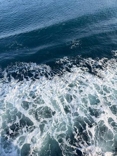 The image provides a detailed, downward-looking perspective of dark blue seawater, illustrating the wake created by a moving vessel. The upper half of the frame shows relatively calm, deep blue water with subtle ripples on its surface. The lower half is dominated by a turbulent area of white, frothy foam and aerated lighter blue water, characteristic of a stern wake from a boat or ship. No individuals, specific vessels, or land formations are visible within the frame. The scene captures the dynamic interaction of water and propulsion, consistent with open water conditions observed in the vicinity of Palaiopoli, Greece.