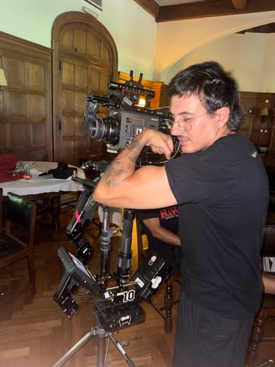 An indoor scene in Hurlingham, Argentina, depicts a male individual and a professional video camera setup. The male, positioned right of center, has dark, medium-length hair, wears round eyeglasses, and a plain black short-sleeve t-shirt. He is actively flexing his heavily tattooed left bicep, with his right hand near his chin, appearing focused on his arm. To his left, a large digital cinema camera, branded "SONY" and equipped with "TILTA" accessories, is mounted on a complex tripod or dolly system. The camera features a large lens, external monitors, and numerous cables, with a "10" marker visible on one accessory.

In the background, the environment consists of rich, dark wooden wall paneling and a prominent arched wooden doorway. A portion of a wooden ceiling structure is visible at the top. To the left, a table covered with a white tablecloth and draped with red fabric holds a small vase with a floral arrangement. Dark upholstered chairs are partially visible around the table and throughout the room. A second individual is partially visible behind and to the right of the primary male subject, wearing a black t-shirt with "RAW" text. The floor is covered with dark hardwood. The lighting appears warm, casting some shadows.