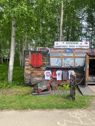 The image depicts an outdoor souvenir stall in Tal'tsy, Russia, nestled amongst birch trees. The stall itself is constructed from weathered wood, resembling a rustic structure with a slightly slanted roof. A large red shield-shaped decoration is affixed to the left side of the stall. In front of the stall, several t-shirts and a tote bag are displayed on hangers. To the left of the t-shirts, there is a wooden object resembling an old cannon, adorned with red accents. A signpost stands in front of the stall, displaying information in Cyrillic script, including the word "coffee". The ground is covered in grass, with scattered yellow wildflowers, and a paved road runs in the foreground. The overall impression is a quaint, forested roadside attraction offering local crafts and refreshments.