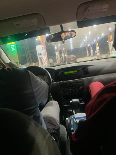 A view from the driver's seat of a car, possibly a Volkswagen, facing a gas station in Jiru, Nigeria. Multiple gas pumps are visible through the windshield. The driver is partially visible, wearing a gray and black jacket. The dashboard features a radio with a green display. The rearview mirror is visible, and the sun visor is down on the driver's side. A red garment is on the passenger side.