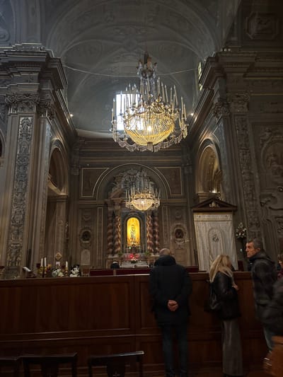The image depicts the interior of the Basilica di San Francesco in Ferrara, Italy. The scene shows a grand, ornate Baroque church nave with a high, dark, vaulted ceiling adorned with faded frescoes. Two large, multi-tiered crystal chandeliers, brightly illuminated, hang prominently from the ceiling, with the upper chandelier catching natural light from an unseen window. The walls feature dark paneling or paint, accented by extensive gilded molding, decorative relief sculptures, and large fluted pilasters with Corinthian capitals.
In the center, a brightly lit side altar or shrine is visible, featuring a golden niche housing a light-colored statue, flanked by twisted, ornate columns. Red fabrics, white flowers, and candle holders are arranged at the base of this shrine, above which hangs a smaller chandelier. To the left of the shrine, a dark wooden stand holds several white candles and flowers. To the right, a tall, narrow structure draped in white fabric with a decorative wooden pediment stands.
In the foreground, a dark wooden partition or pews are present. Three individuals are visible: one male, center-left, seen from the back, wearing a dark winter coat and trousers, standing with hands clasped behind his back, observing the shrine; one female, center-right, with long blonde hair, wearing a dark jacket and a light-colored long skirt, also observing the shrine; and one male, far right, in profile, with short, curly dark hair and a dark patterned jacket, looking towards the right side of the image. Indistinct figures are visible further in the background near the central shrine. The overall atmosphere is quiet and observant.