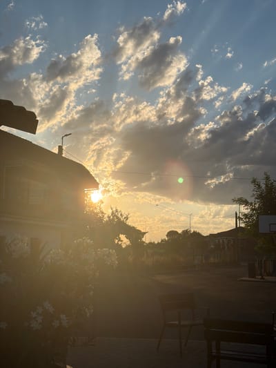 This image captures a serene sunset scene in Arcahueja, Spain. The sky is a gradient of blue and warm hues, illuminated by the setting sun peeking through scattered clouds. Sunlight streams down, creating a lens flare effect and casting a warm glow over the environment. In the foreground, a residential building with a dark roof is partially visible on the left, with white flowers in bloom in front of it. The middle ground shows a street or courtyard with two dark benches and a basketball hoop in the distance. Several trees are scattered throughout the scene. The overall atmosphere is peaceful and tranquil.