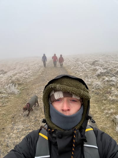 The image depicts a group of four individuals and a dog hiking on a frosty, unpaved path through moorland in West Devon, United Kingdom, consistent with terrain found in Dartmoor National Park. In the foreground, a person is taking a selfie, visible from the chest up. This individual wears a black hooded jacket with a dark green fleece-lined hood, a light brown and cream checkered knitted hat, and a grey fleece neck gaiter covering their mouth and nose. A nose piercing is visible on the right nostril. A grey and yellow backpack strap is visible on their right shoulder. To the left of this person, a medium-brown dog, wearing a khaki-green dog coat, walks on the path with its head down. Further along the path, three additional individuals are observed walking in a single file away from the camera. The lead person wears a blue jacket, followed by two individuals wearing red jackets; the middle person holds a walking stick in their right hand. The ground is covered with short, sparse vegetation, appearing light brown and white due to frost. Dense fog significantly reduces visibility, obscuring the horizon and creating a pale, indistinct background. The scene suggests cold weather conditions.