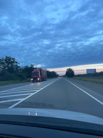 The image depicts a highway scene at dusk. A red truck with a cargo container is traveling on the right lane, with its headlights illuminated. The highway has white lane markings, and the surrounding environment consists of trees and vegetation on the left and a field on the right. In the distance, other vehicles can be seen, and a billboard stands in the field. The sky is filled with dramatic clouds in shades of blue, with hints of pink and orange near the horizon. The foreground shows the dashboard and windshield of a vehicle. The location is identified as the highway near Abinsk, Russia.